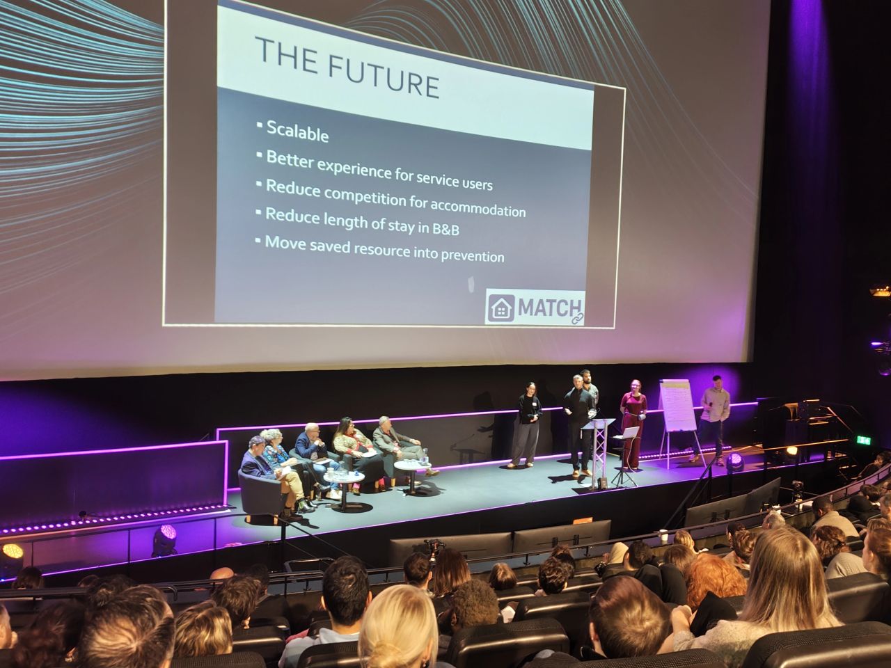 Becki Floyd presenting MATCH on stage, with the MATCH slide behind her